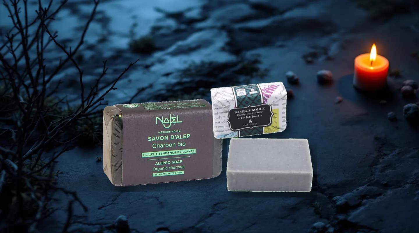 Aleppo Seife mit Holzkohle von Najel in schwarzer Banderole und Thai Naturseife Bambus Charcoal mit Zeitungspapier-Verpackung auf dunklem Schiefer neben kahlen Zweigen und einer Kerze